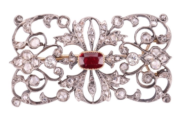 A late Victorian diamond and ruby brooch, of scrolled foliate design, the openwork mount encrusted with old-cut diamonds and rose-cut diamond points, centred with a cushion-cut ruby in collet with twisted cable edge, measures 6.7 x 4.7 mm, unmarked metal 