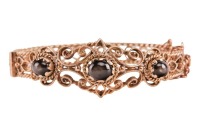 A hinged bracelet set with black star sapphires; the openwork scrolled mount with twisted rope border and beaded accents, set with three graduated oval black star sapphire cabochons, fastens with a tongue clasp with push-in thumbpiece, in yellow metal sta