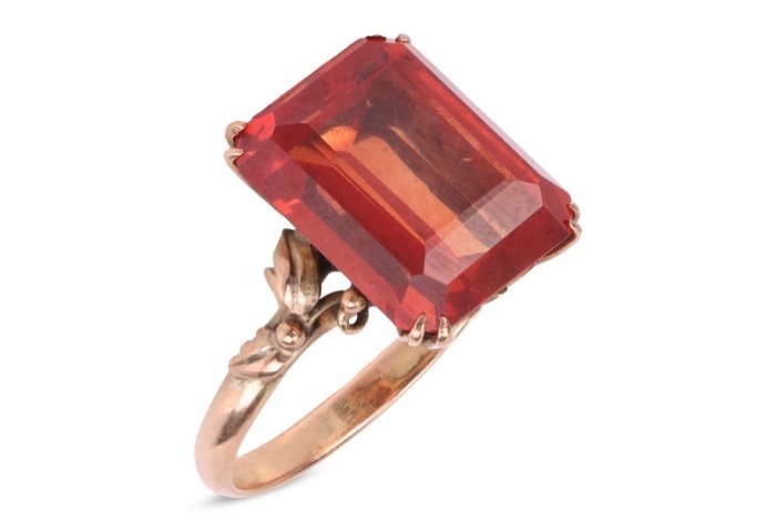 A synthetic orange sapphire ring, with an emerald-cut synthetic sapphire in reddish-orange colour, double claw set on a yellow metal mount, floral details on gallery and shoulders, to a D-profile shank, marked '14k', ring size O, total weight of item 4.8 