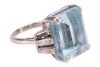 An Art Deco style aquamarine and diamond-set ring, claw-set with a step-cut aquamarine, measuring approximately 15.8mm x 13.5mm, flanked by six baguette-cut diamonds, in a tapering design, to an unmarked white metal shank surface tested as 18ct; 10.0 gram - 3