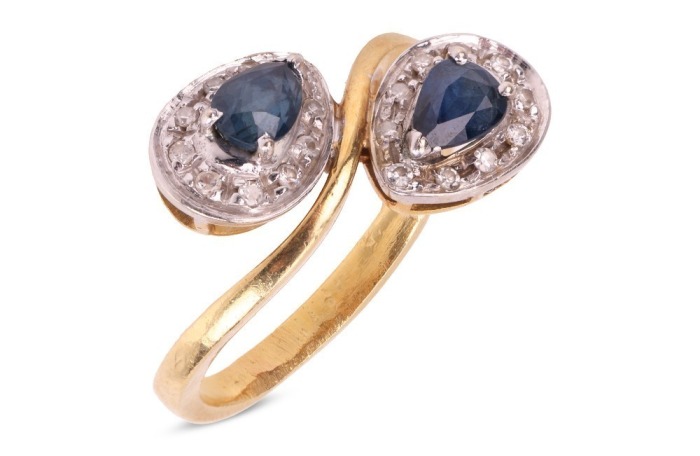 A sapphire and diamond crossover ring, consisting of two pear-shaped sapphires claw-set on white metal mounts, each encircled with diamond accents, to a tapered flat shank in yellow metal, marked as '750', ring size M&frac12;, total weight of item 4.6 grams.