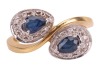 A sapphire and diamond crossover ring, consisting of two pear-shaped sapphires claw-set on white metal mounts, each encircled with diamond accents, to a tapered flat shank in yellow metal, marked as '750', ring size M&frac12;, total weight of item 4.6 grams. - 2