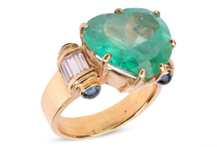 A large heart-shaped emerald dress ring, the emerald measures 13.8 x 15.0 x 8.2 mm, claw-set to the centre between trumpeted shoulders of scroll form, embellished with baguette-cut diamonds and sapphire cabochon terminals, leading to a tapering flat shank
