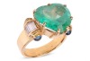 A large heart-shaped emerald dress ring, the emerald measures 13.8 x 15.0 x 8.2 mm, claw-set to the centre between trumpeted shoulders of scroll form, embellished with baguette-cut diamonds and sapphire cabochon terminals, leading to a tapering flat shank